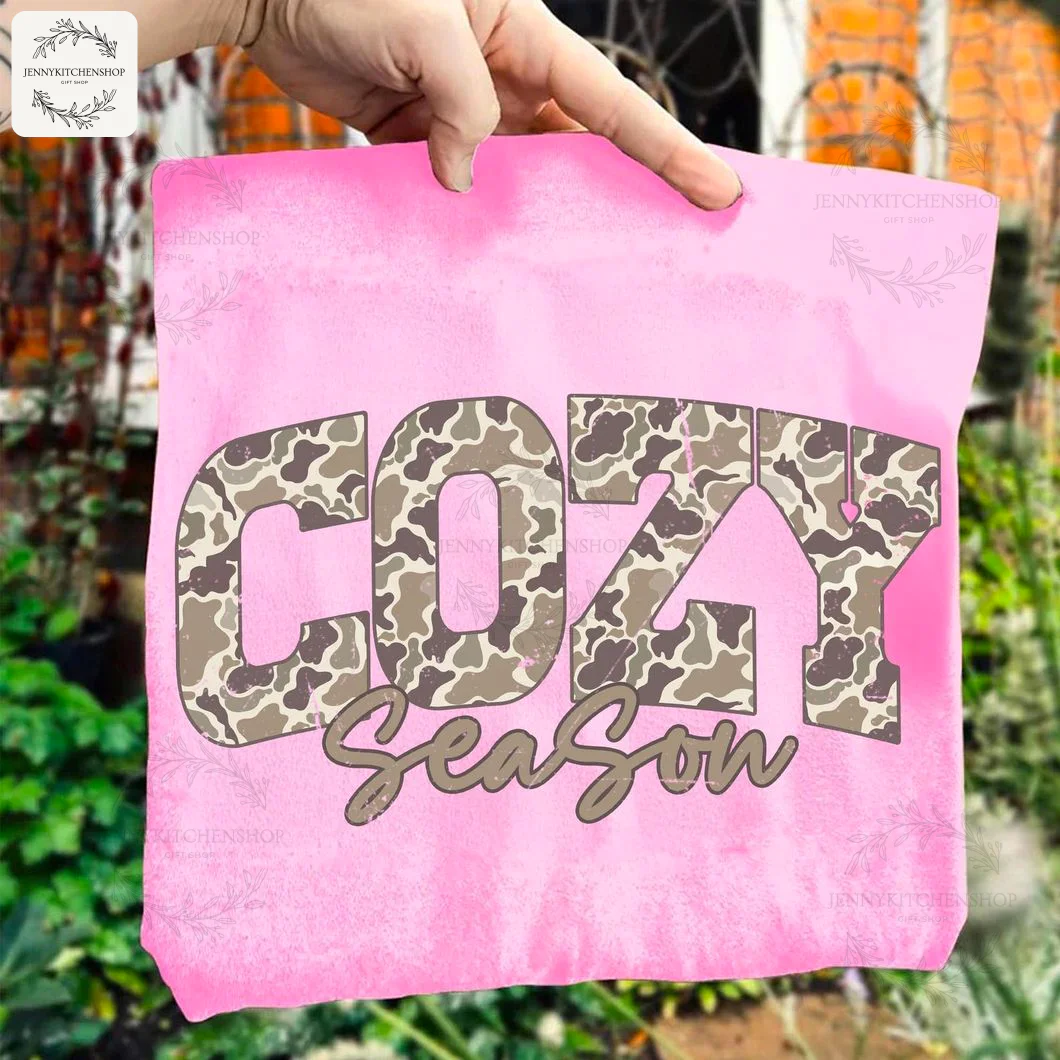 A hand holds up a bright pink t-shirt to showcase the "Cozy Season" camouflage sublimation print design close up.