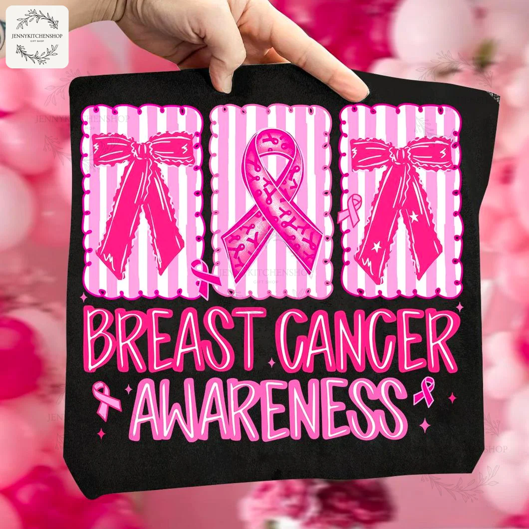 A hand holds up a black t-shirt to show the vibrant pink coquette design for breast cancer awareness against a blurry pink background.