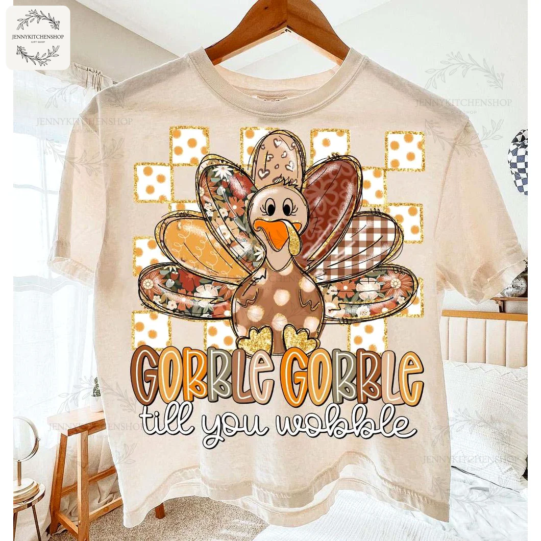 A cream-colored Thanksgiving t-shirt with a cute patchwork turkey and the phrase "Gobble Gobble till you wobble" hanging on a hanger.